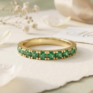 May include: A gold eternity band ring featuring a continuous row of round-cut, vibrant green gemstones set in a shared-prong setting. The ring has a polished metallic finish and a uniform circular shape, showcasing a classic design with bright stones.