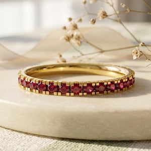 May include: A gold eternity band featuring a continuous row of small, round, deep red rubies set in a channel setting. The ring has a polished metallic finish and a uniform circular shape, showcasing a vibrant contrast between the gold and red.