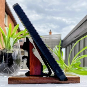 Puede incluir: Un soporte de teléfono de madera con una silueta de gato negro y una incrustación de madera roja y marrón. Sostiene un teléfono negro, sobre un fondo de cielo nublado. La base es de madera marrón oscuro.
