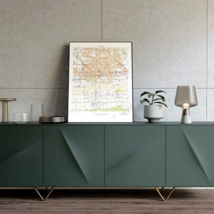 May include: A dark green modern sideboard with gold-colored legs. The sideboard has four doors and a geometric design. On top are decorative items: a framed map, a small lamp, a potted plant, and other decorative objects.
