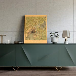 May include: A dark green modern sideboard with gold legs, featuring a framed vintage map, a small potted plant, a table lamp, and decorative items. The sideboard has a geometric design on the doors.