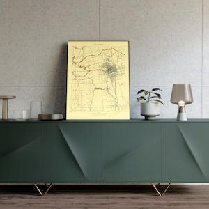 May include: A dark green sideboard with gold legs, displaying a framed vintage map, a small potted plant, and a decorative lamp. The map is in a black frame with a beige background. The sideboard is against a light grey tiled wall.