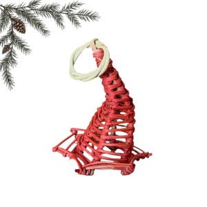 Pode incluir: Um ornamento de vime trançado vermelho em forma de chapéu pontiagudo com aba larga e um laço circular bege claro no topo. A estrutura é feita de fibras naturais finas entrelaçadas, criando um design de treliça aberto e vazado.