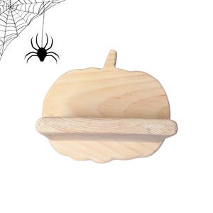 Pode incluir: Um suporte de madeira de cor clara em forma de abóbora com uma ranhura horizontal no centro. Um gráfico preto de uma aranha pendurada numa teia está posicionado no canto superior esquerdo, separado do objeto de madeira.