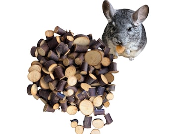 Könnte beinhalten: Ein graues Chinchilla mit großen Ohren und Schnurrhaaren hält ein kleines, rundes Kauspielzeug aus hellem Holz. Neben dem Chinchilla liegt ein großer Haufen kleiner, zylindrischer Holzstücke mit dunkler Rinde und hellen, runden Schnittflächen.