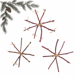 Pode incluir: Três ornamentos em forma de floco de neve feitos de galhos finos castanho-avermelhados amarrados com barbante castanho-claro. Cada ornamento apresenta um centro com ramos radiantes, acompanhado por um pequeno ramo de pinheiro verde com pinhas.