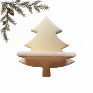 Pode incluir: Uma prateleira de parede de madeira de cor clara em forma de árvore perene estilizada com uma superfície horizontal plana para exibição. A prateleira apresenta um acabamento liso de grão de madeira natural e um ramo de pinheiro decorativo.