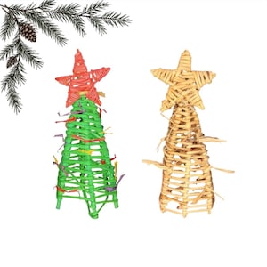 Pode incluir: Dois ornamentos em forma de árvore cónica feitos de fibras vegetais entrançadas. Uma árvore é verde brilhante com uma estrela vermelha no topo e tiras de papel coloridas, a outra é castanha natural com uma estrela a condizer.