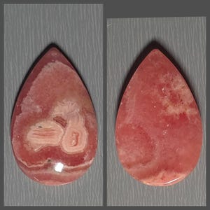 Pode incluir: Dois cabochões de rodocrosita em forma de gota com superfície polida. As pedras exibem uma paleta de cores rosa vibrante e salmão com distintos padrões de faixas concêntricas brancas e rosa claro e inclusões minerais naturais.