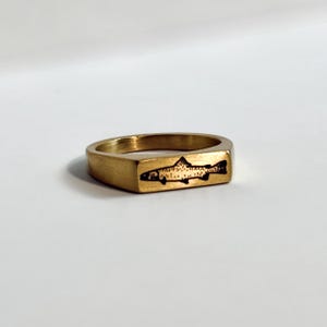 May include: A gold-colored ring with a rectangular face. The ring features a black fish design etched into the face. The ring is set against a white background.