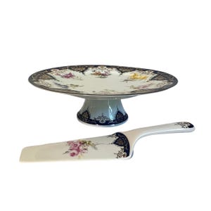 May include: A white porcelain cake stand and server set. The cake stand has a round plate with a dark blue and gold border, supported by a pedestal base. The server features a floral design with pink roses and a matching blue and gold trim.