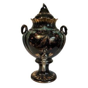 May include: A black ceramic samovar with gold accents and floral designs. The antique tea kettle features a spigot, two handles, and a decorative lid. The base is round and ornate, and the overall design is vintage.