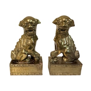 May include: A pair of brass Foo Dog statues. Each dog is seated on a rectangular base with a detailed, patterned border. The dogs have a fierce expression and detailed fur. The statues are a shiny gold color.