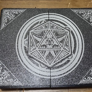 May include: A black, hinged box with a silver geometric design. The design features a central dodecahedron within a circle, with ornate corner patterns. The box is likely for storing dice or other gaming accessories.