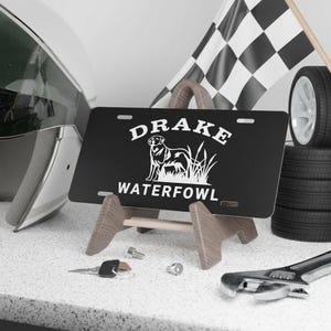 May include: Black license plate with the word "DRAKE" above a dog illustration and the word "WATERFOWL" below. The plate is displayed on a wooden stand, with a checkered flag and tires in the background.