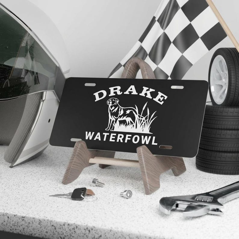 May include: Black license plate with the word "DRAKE" above a dog illustration and the word "WATERFOWL" below. The plate is displayed on a wooden stand, with a checkered flag and tires in the background.