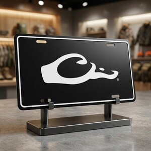 May include: Black license plate with a white logo of a duck head. The license plate is displayed on a metal stand. The license plate has a white border and is displayed on a gray surface.
