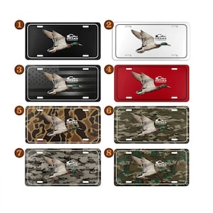 May include: A collection of eight license plates featuring a flying duck graphic. The plates have various backgrounds, including black, white, red, camouflage, and an American flag design. Each plate includes the "DRAKE" logo.