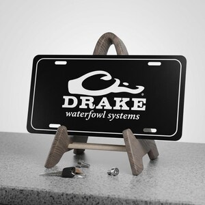 May include: Black license plate with white border and text "DRAKE waterfowl systems." The plate is displayed on a wooden easel. A set of keys, bolts, and a small metal object are in front of the easel.