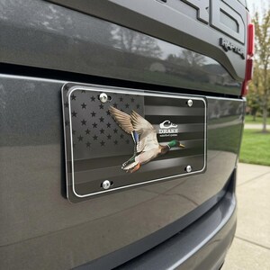 May include: License plate featuring a flying mallard duck over a black and gray American flag design. The plate includes the text "DRAKE waterfowl systems" and is mounted on a gray vehicle.