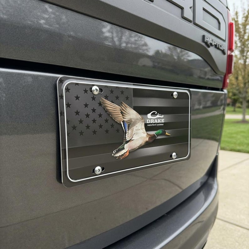 May include: License plate featuring a flying mallard duck over a black and gray American flag design. The plate includes the text "DRAKE waterfowl systems" and is mounted on a gray vehicle.