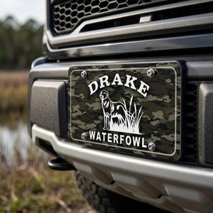 May include: License plate with a camouflage pattern and the text "DRAKE WATERFOWL" in white. A white silhouette of a dog and tall grass is centered on the plate. The plate is mounted on the front of a black truck.