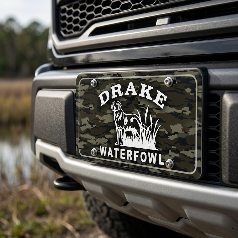 May include: License plate with a camouflage pattern and the text "DRAKE WATERFOWL" in white. A white silhouette of a dog and tall grass is centered on the plate. The plate is mounted on the front of a black truck.