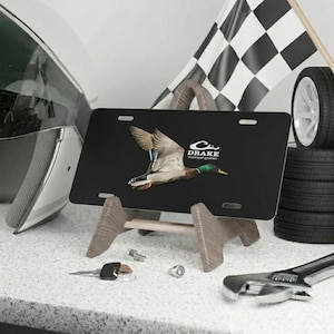 May include: Black license plate with a flying duck graphic and the text "DRAKE waterfowl games." The plate is displayed on a wooden stand, with a checkered flag and a tire in the background.