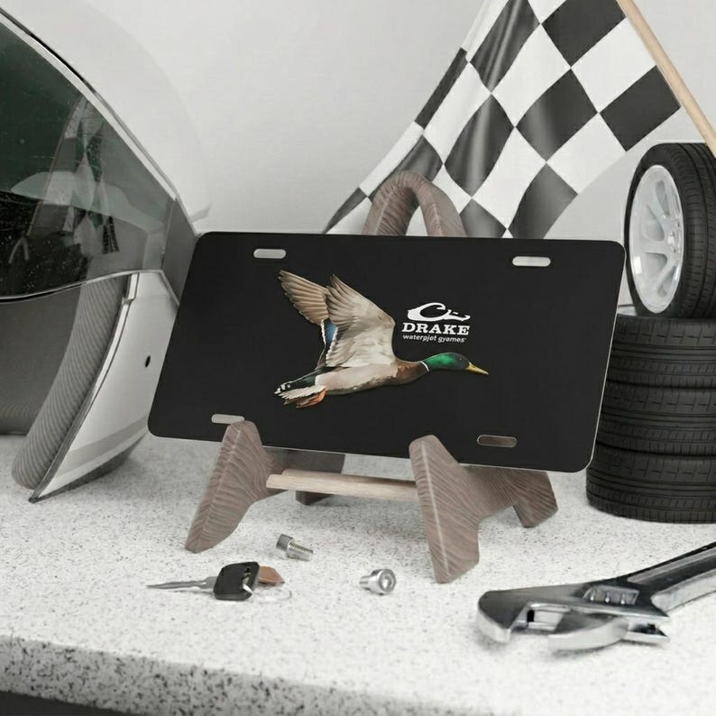 May include: Black license plate with a flying duck graphic and the text "DRAKE waterfowl games." The plate is displayed on a wooden stand, with a checkered flag and a tire in the background.