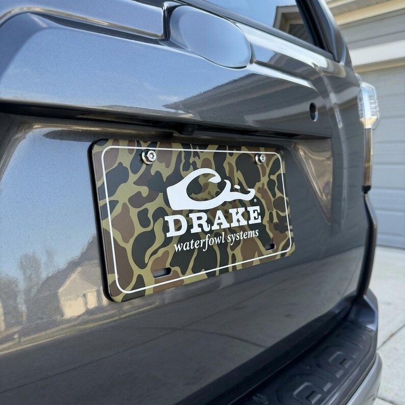 May include: A license plate with a camouflage pattern and the text "DRAKE waterfowl systems" in white. The plate is mounted on a gray vehicle.