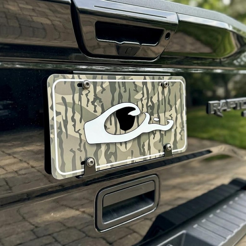 May include: License plate with a camouflage pattern and a white graphic of a duck head and the word "C". The plate is mounted on a black vehicle. The plate is held in place with four silver screws.