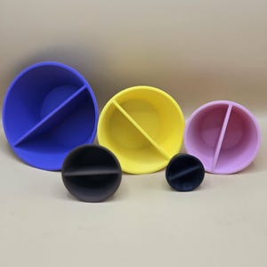May include: A collection of colorful, divided bowls and matching black, round objects. The bowls are in blue, yellow, and pink, each with a divider creating two compartments. The black objects are also divided, with a line across the middle.