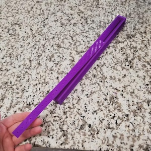 May include: A purple, rectangular tool with a series of small holes along one side. The tool is made of a solid material and has a channel running along its length. The tool is held by a hand.