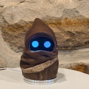 May include: A brown hooded figure figurine with two glowing circular blue eyes. The figure wears a textured brown robe and a utility belt across the chest. It is mounted on a circular silver base and features a smooth, matte finish throughout.