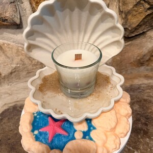 May include: A clear glass candle with a wooden wick sits inside a cream-colored, open seashell-shaped holder. The base features a blue pool with a bright pink starfish, surrounded by textured, light orange rock-like formations on a white stand.