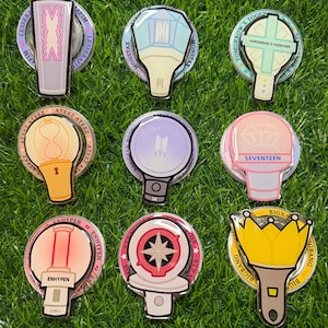 May include: Nine colorful K-pop light stick pins on a green grass background. Each pin features a unique design and logo, including groups like BTS, Seventeen, and Bigbang. The pins are round with a clear, glossy finish.