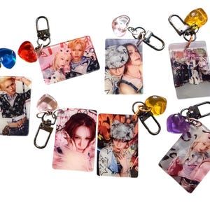 May include: A collection of rectangular keychains featuring images of people, each attached to a metal clasp. The keychains have various colored heart-shaped and gem-like charms. The images on the keychains are of people.