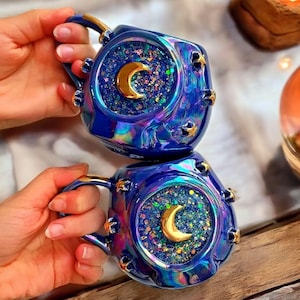 Pode incluir: Duas canecas de cerâmica azul iridescente com detalhes dourados. Cada caneca apresenta um design de lua crescente e estrelas, com glitter no interior do círculo. As alças e as estrelas são douradas.