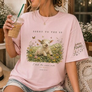 May include: A light pink t-shirt featuring a graphic of a smiling lamb in a field of wildflowers with butterflies. The text reads SORRY TO THE 99, I took the scenic route, LURE 15:4, and LOST FOUND on the sleeve. The shirt has a crew neckline.