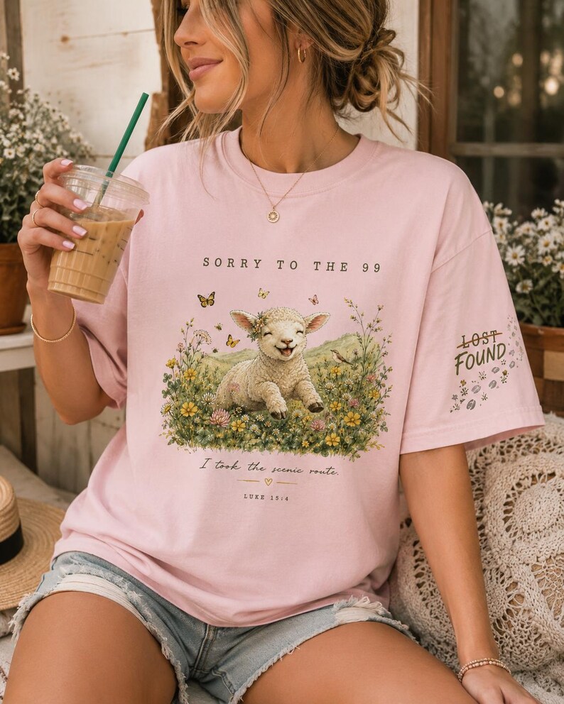 May include: A light pink t-shirt featuring a graphic of a smiling lamb in a field of wildflowers with butterflies. The text reads SORRY TO THE 99, I took the scenic route, LURE 15:4, and LOST FOUND on the sleeve. The shirt has a crew neckline.