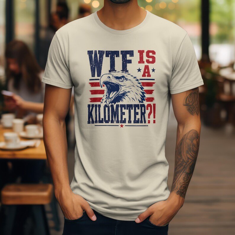 Pode incluir: Camiseta bege clara com um design gr&aacute;fico. O design apresenta uma &aacute;guia patri&oacute;tica e o texto "WTF IS A KILOMETER?!" em azul marinho e vermelho. O design inclui estrelas e listras.