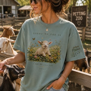 May include: A sage green short-sleeve t-shirt featuring a graphic of a lamb in a field of flowers with butterflies. The text reads SORRY TO THE 99, LOST FOUND, I took the scenic route, and LUKE 15:4. The shirt has a crew neck and a relaxed fit.