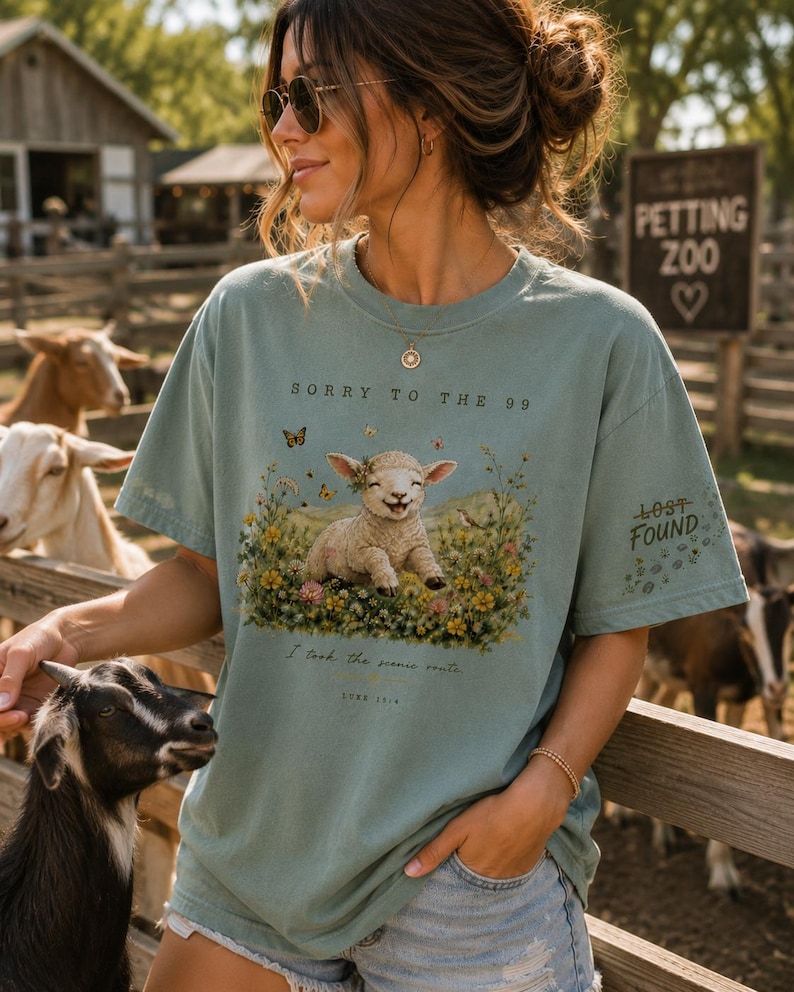 May include: A sage green short-sleeve t-shirt featuring a graphic of a lamb in a field of flowers with butterflies. The text reads SORRY TO THE 99, LOST FOUND, I took the scenic route, and LUKE 15:4. The shirt has a crew neck and a relaxed fit.