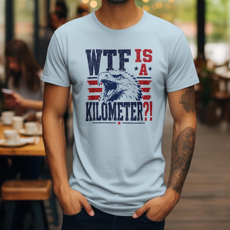 Pode incluir: Camiseta azul clara com um gr&aacute;fico de uma &aacute;guia e o texto "WTF IS A KILOMETER?!" em azul marinho e vermelho. O design inclui estrelas e listras, dando-lhe um tema patri&oacute;tico. A camiseta &eacute; feita de um material macio.