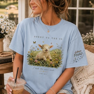 May include: A light blue t-shirt featuring a graphic of a smiling lamb sitting in a field of colorful wildflowers and butterflies. The text reads SORRY TO THE 99, I took the scenic route, LUKE 15:4, and LOST FOUND on the sleeve with small floral accents.