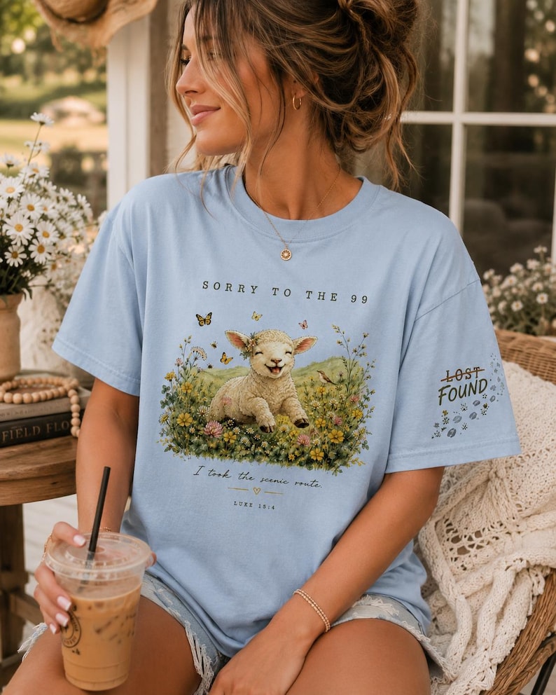 May include: A light blue t-shirt featuring a graphic of a smiling lamb sitting in a field of colorful wildflowers and butterflies. The text reads SORRY TO THE 99, I took the scenic route, LUKE 15:4, and LOST FOUND on the sleeve with small floral accents.