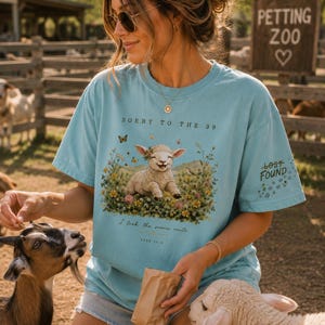 May include: A light blue t-shirt featuring a graphic of a lamb sitting in a field of colorful wildflowers. The text reads SORRY TO THE 99, I took the scenic route, LUKE 15:4, and LOST FOUND on the sleeve. The shirt has a crew neck and short sleeves.