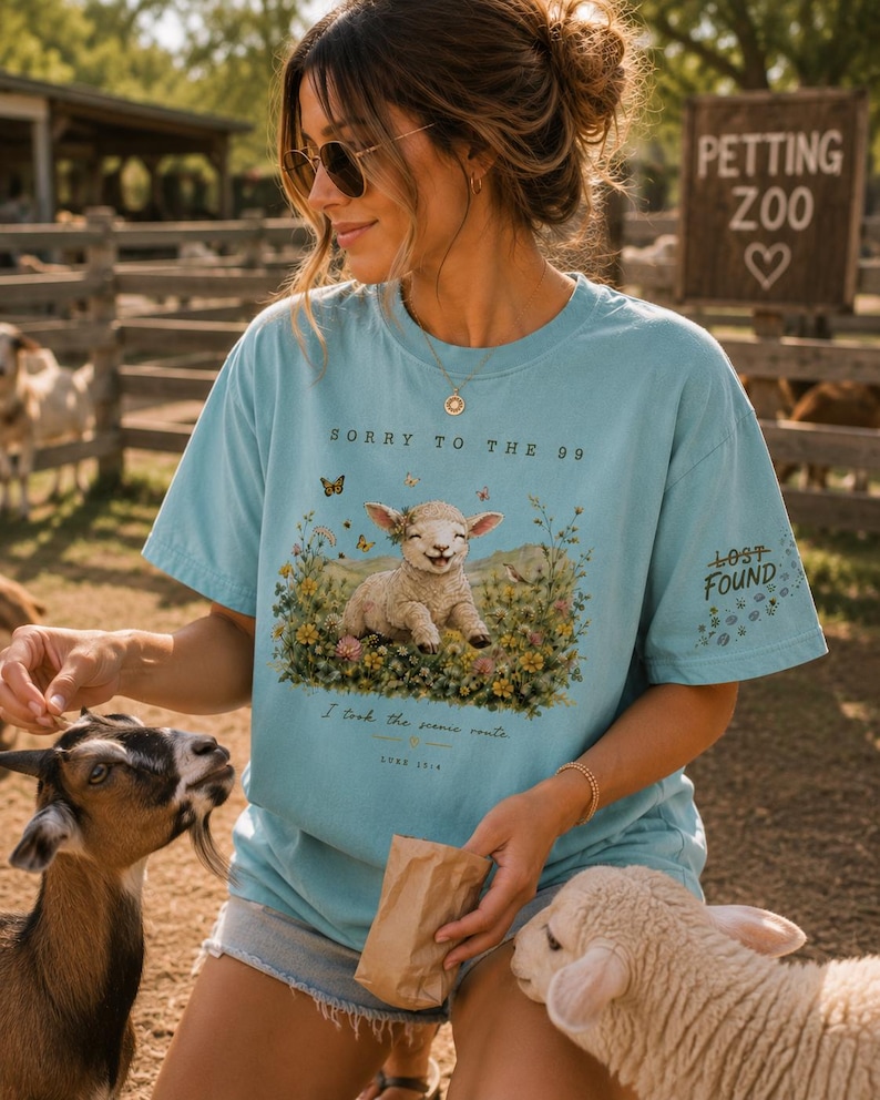 May include: A light blue t-shirt featuring a graphic of a lamb sitting in a field of colorful wildflowers. The text reads SORRY TO THE 99, I took the scenic route, LUKE 15:4, and LOST FOUND on the sleeve. The shirt has a crew neck and short sleeves.