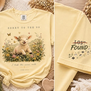 May include: A pale yellow t-shirt featuring a graphic of a smiling lamb in a field of wildflowers with butterflies. The text reads SORRY TO THE 99, I took the scenic route, and LUKE 15:4. The sleeve displays the text LOST FOUND with small hoof prints.