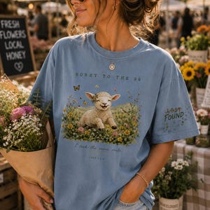 May include: A light blue t-shirt featuring a graphic of a smiling lamb sitting in a field of colorful wildflowers and butterflies. The text reads Sorry to the 99, Lost Found, I took the scenic route, and Luke 15:4. The shirt has a crew neckline.